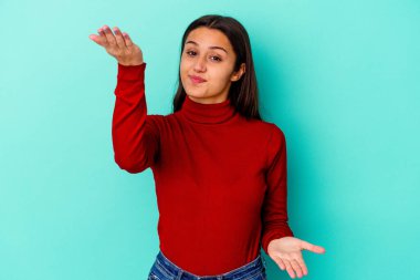 Mavi arka planda izole edilmiş genç Hintli kadın kollarıyla ölçeği yapıyor, mutlu ve kendinden emin hissediyor..