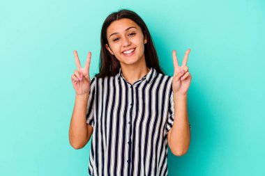 Mavi arka planda izole edilmiş genç Hintli kadın zafer işareti gösteriyor ve gülümsüyor..