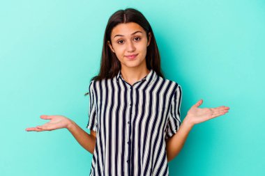 Genç Hintli bir kadın, mavi arka planda izole edilmiş şüphe içinde ve sorgulama sırasında omuz silkiyor..