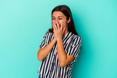 Mavi arka planda izole edilmiş genç Hintli bir kadın bir şeye gülüyor, elleriyle ağzını kapatıyor..