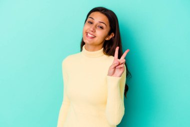 Mavi arka planda izole edilmiş genç Hintli kadın neşeli ve kaygısız parmakları olan bir barış sembolü gösteriyor..