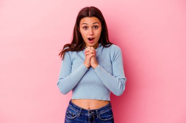 Pembe arka planda izole edilmiş genç Hintli bir kadın şans için dua ediyor, şaşırıyor ve ağzını açıyor..