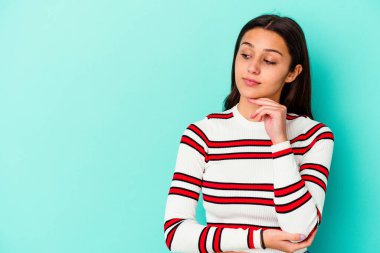 Mavi arka planda izole edilmiş genç Hintli kadın şüpheli ve şüpheci bir ifadeyle yana bakıyor..