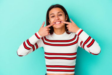 Mavi arka planda izole edilmiş genç Hintli kadın gülümsüyor, parmaklarını ağıza işaret ediyor..