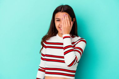 Mavi arka planda izole edilmiş genç Hintli kadın yüzünün yarısını avuç içiyle kaplayarak eğleniyor..