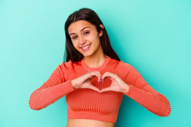 Mavi arka planda izole edilmiş genç Hintli kadın gülümsüyor ve elleriyle kalp şeklini gösteriyor..