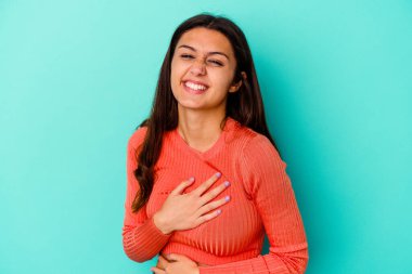 Mavi arka planda izole edilmiş genç Hintli kadın mutlu bir şekilde gülüyor ve ellerini karnının üstünde tutarak eğleniyor..