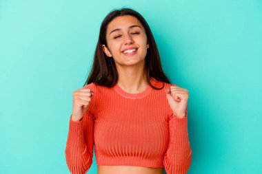 Genç Hintli kadın mavi arka planda izole edilmiş zafer, tutku ve coşkuyu kutluyor, mutlu bir ifade..