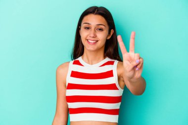 Mavi arka planda izole edilmiş genç Hintli kadın zafer işareti gösteriyor ve gülümsüyor..