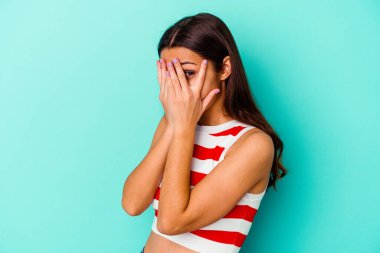 Mavi arka planda izole edilmiş genç Hintli kadın parmaklarının arasından korkmuş ve gergin bir şekilde göz kırpıyor..