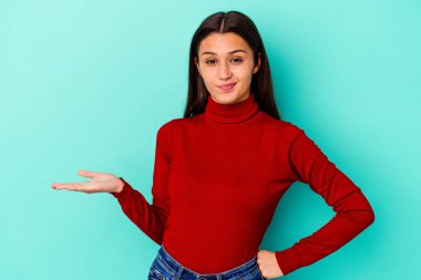 Genç Hintli bir kadın, mavi arka planda izole edilmiş şüphe içinde ve sorgulama sırasında omuz silkiyor..