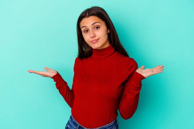 Mavi arka planda izole edilmiş genç bir Hintli kadın. Kafası karışmış ve omuz silkiyor. Kopya alanını tutmak için..