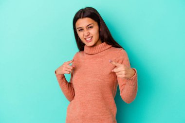 Mavi arka planda izole edilmiş genç Hintli kadın parmaklarıyla ön tarafı işaret ediyor..