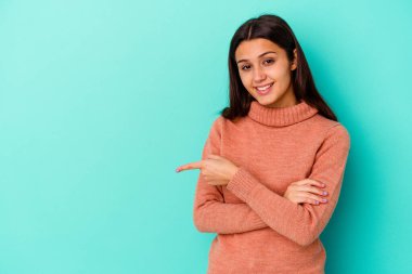 Mavi arka planda izole edilmiş genç Hintli kadın işaret parmağıyla işaret ederek gülümsüyor..