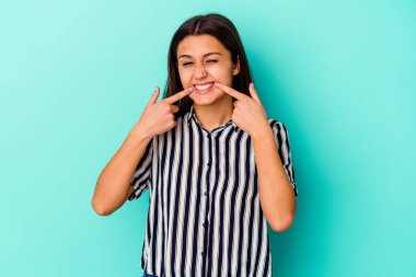 Mavi arka planda izole edilmiş genç Hintli kadın gülümsüyor, parmaklarını ağıza işaret ediyor..