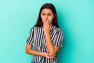 Mavi arka planda izole edilmiş genç bir Hintli kadın esniyor. Ağzı eliyle kapatan yorgun bir hareket sergiliyor..