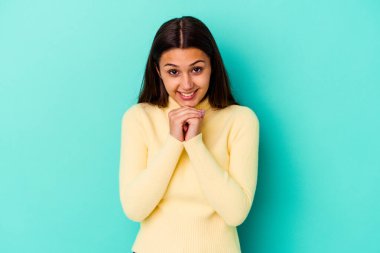 Mavi arka planda izole edilmiş genç Hintli kadın ellerini çenesinin altında tutuyor ve mutlu bir şekilde yan tarafa bakıyor..