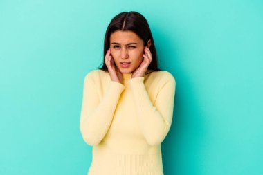 Genç Hintli bir kadın mavi arka planda izole edilmiş kulakları parmaklarıyla kapatıyor, stresli ve çaresiz bir şekilde yüksek sesli bir ortam tarafından.