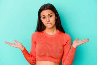 Mavi arka planda izole edilmiş genç bir Hintli kadın. Kafası karışmış ve omuz silkiyor. Kopya alanını tutmak için..