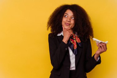Genç Afro-Amerikan hostes elinde küçük bir uçak tutuyor sarı arka planda izole edilmiş şüpheli ve şüpheci bir ifadeyle yana bakıyor..