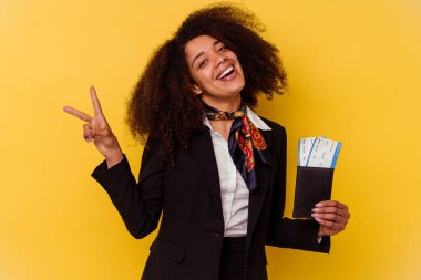 Genç Afrikalı Amerikalı hostes sarı arka planda izole edilmiş bir uçak bileti tutuyor neşeli ve tasasız parmakları olan bir barış sembolü gösteriyor..