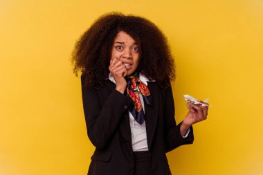 Genç Afrikalı Amerikalı hostes sarı arka planda izole edilmiş, tırnak yiyen, gergin ve endişeli bir uçak tutuyor..
