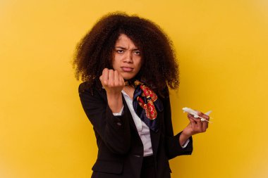 Genç Afrikalı Amerikalı hostes, sarı arka planda izole edilmiş küçük bir uçak tutuyor. Kameraya yumruk, agresif yüz ifadesi gösteriyor..