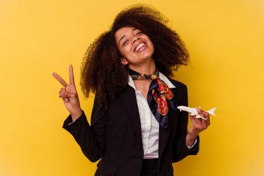 Genç Afrikalı Amerikalı hostes sarı arka planda izole edilmiş küçük bir uçak tutuyor neşeli ve tasasız parmakları olan bir barış sembolü gösteriyor..