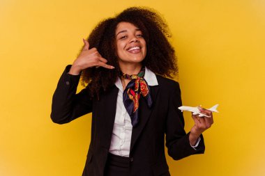 Genç Afrikalı Amerikalı hostes sarı arka planda izole edilmiş küçük bir uçak tutuyor. Parmakları olan bir cep telefonu hareketini gösteriyor..