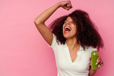 Genç Afrikalı Amerikalı kadın pembe arka planda izole edilmiş sağlıklı bir smoothie içiyor zafer kazandıktan sonra yumruk kaldırıyor, kazanan konsept.
