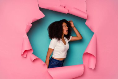 Kırık kağıda sarılı genç Afrikalı Amerikalı kadın mavi arka planda izole edilmiş, uzaklara bakıyor ve elini alnına koyuyor..