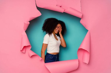 Kırık kağıda sarılı genç Afrikalı Amerikalı kadın mavi arka planda izole edilmiş parmaklarıyla kameraya göz kırpıyor, utanarak yüzünü kapatıyor..