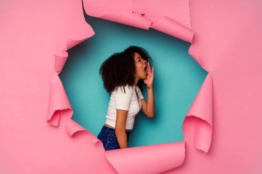 Kırık kağıda sarılmış genç Afrikalı Amerikalı kadın mavi arka planda izole edilmiş bağırıyor ve avuç içi açık bir şekilde ağzını tutuyor..