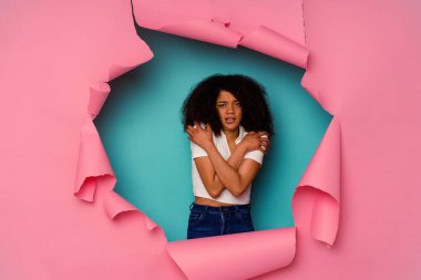 Kırık kağıda sarılı genç Afrikalı bir kadın mavi arka planda izole edilmiş. Düşük ateş ya da hastalık nedeniyle soğuyor..