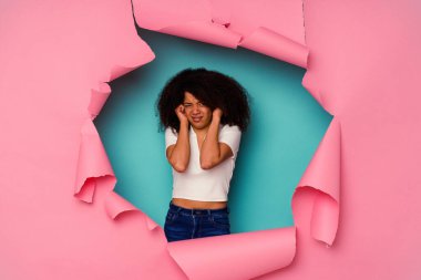 Kırık kağıda sarılı genç Afrikalı Amerikalı kadın mavi arka planda izole edilmiş kulakları elleriyle kapatıyor..