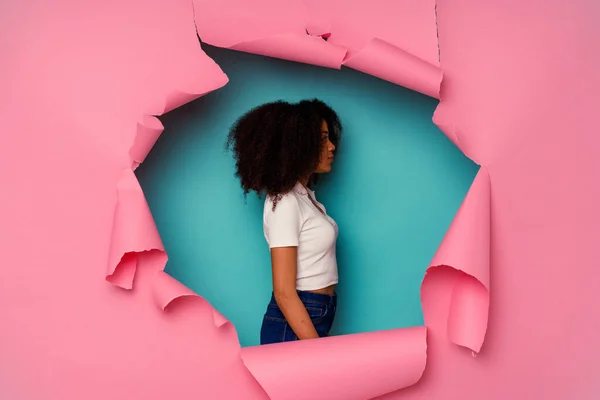 Young African American woman in torn paper isolated on blue background gazing left, sideways pose.