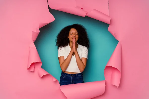 Young African American woman in torn paper isolated on blue background holding hands in pray near mouth, feels confident.