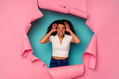 Genç Afrikalı Amerikalı bir kadın yırtık bir kağıtta mavi arka planda izole edilmiş bir şekilde bir başarı fırsatı bulmak için gözlerini açık tutuyor..