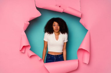 Mavi arka planda izole edilmiş yırtık bir kağıda sarılmış genç Afrikalı Amerikalı kadın omuzlarını silker ve gözleri açık kalır..