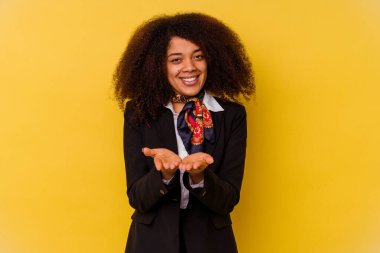 Genç Afrikalı Amerikalı hostes sarı arka planda izole edilmiş. Elinde avuç içi olan bir şeyle kameraya sunuyor..