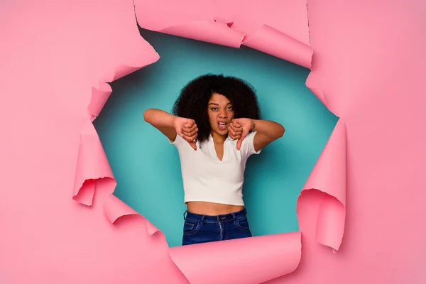 Young African American woman in torn paper isolated on blue background showing thumb down and expressing dislike.