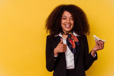 Genç Afrikalı Amerikalı hostes sarı arka planda izole edilmiş küçük bir uçak tutuyor gülümsüyor ve başparmağını kaldırıyor.