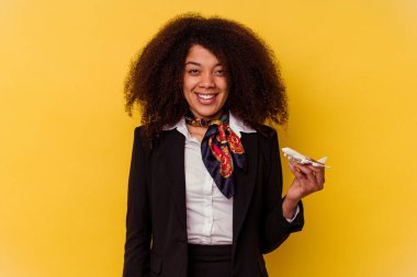 Genç Afro-Amerikan hostes elinde küçük bir uçak tutuyor. Sarı arka planda izole edilmiş. Mutlu, gülümseyen ve neşeli..