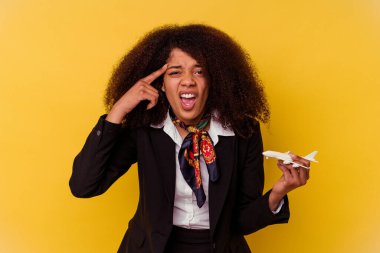 Genç Afro-Amerikan hostes sarı arka planda izole edilmiş küçük bir uçak tutuyor işaret parmağıyla hayal kırıklığı jesti gösteriyor..