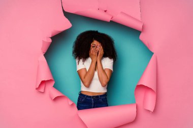 Kırık kağıda sarılı genç Afrikalı Amerikalı kadın mavi arka planda izole edilmiş. Korkmuş ve gergin parmaklarıyla göz kırp..