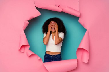Mavi arka planda izole edilmiş yırtık bir kağıda sarılı genç Afrikalı Amerikalı kadın elleriyle gözlerini kapatıyor, büyük bir gülümsemeyle bir sürpriz bekliyor..