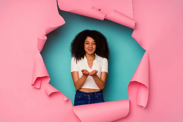 Young African American woman in torn paper isolated on blue background holding something with palms, offering to camera.