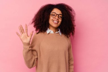 Genç Afrikalı Amerikalı kadın pembe arka planda izole edilmiş. Gülümsüyor. Beş numarayı parmaklarıyla gösteriyor..