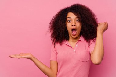 Pembe arka planda izole edilmiş genç Afrikalı Amerikalı kadın avuç içinde fotokopi alanı tutuyor, el üstünde tutuyor. Şaşırmış ve mutlu..