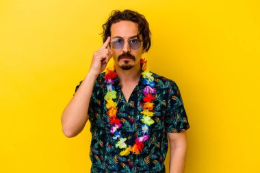 Sarı arka planda izole edilmiş beyaz bir kolye takan genç beyaz bir adam şakağı parmağıyla işaret ediyor, düşünüyor ve bir göreve odaklanıyor..